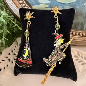 May include: A pair of mismatched gold-tone earrings featuring a star-shaped stud. One earring displays a black witch hat with a yellow crescent moon and red rhinestones. The other earring depicts a witch in a black dress riding a gold broomstick.