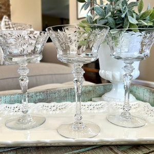 May include: Three clear glass stemmed cocktail glasses with intricate etched floral designs. The glasses are on a white tray with a decorative edge. The tray is on a wooden surface.