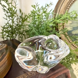 May include: A clear glass rabbit figurine with a rounded body and visible details. The figurine is resting on a dark brown surface, with green foliage and a gold-framed picture in the background. The glass reflects light, creating a shimmering effect.