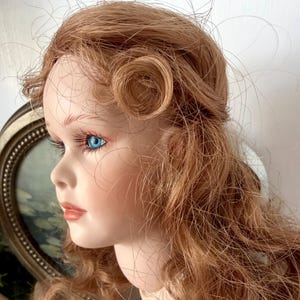 May include: A porcelain doll with long, auburn hair styled with a curled section. The doll has blue eyes, long eyelashes, and a delicate complexion. A framed picture is in the background.
