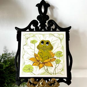 May include: A decorative trivet featuring a cartoon frog on a lily pad. The frog is green and yellow, set against a white background with green leaves. The trivet is framed in black and displayed on a gold-colored stand.