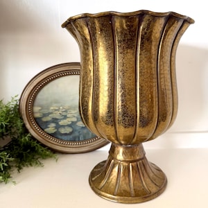 May include: A metallic gold pedestal vase with a scalloped rim and vertical fluted detailing. The surface features a textured, mottled finish throughout the body and the circular base, creating a reflective appearance with dark highlights.