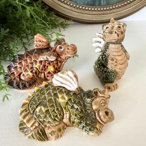 May include: Three ceramic dragon figurines in shades of brown and green. One dragon is standing, while the other two are lying down. The dragons have detailed scales and wings, and are likely decorative items.