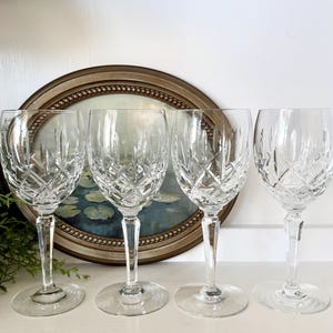 May include: Four clear crystal wine glasses with diamond-cut patterns. The glasses have long stems and round bases. They are arranged in a row in front of a framed painting with a gold-colored frame.