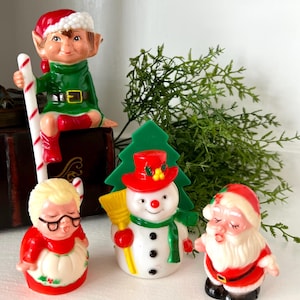 May include: A set of four vintage Christmas figurines: a green elf holding a candy cane, a red and white snowman with a broom, a red and white Santa Claus, and a red and white grandmother figure.