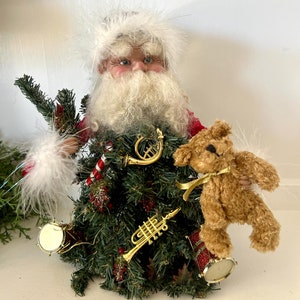 May include: A festive Christmas tree decoration featuring a Santa Claus figure with a white beard and a red hat, holding a teddy bear. The tree is decorated with gold musical instruments, candy canes, and red and white ornaments.