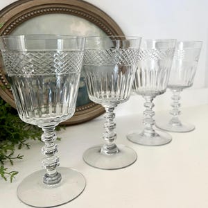 May include: Four clear crystal wine glasses with intricate diamond and vertical line patterns. The stems have a decorative, stacked bead design, and the glasses sit on round bases. The background includes a framed picture and greenery.