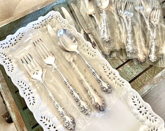 May include: A white ceramic tray with a lace pattern design, holding a set of silver flatware, including forks, spoons, and knives. The flatware is wrapped in clear plastic and arranged neatly on the tray.