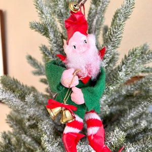 May include: A vintage Christmas ornament featuring a green-clad elf with a red hat and white beard, holding two gold bells with red ribbons. The elf is sitting on a snowy green tree branch.