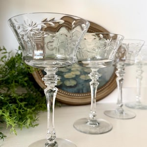 May include: Set of four clear champagne coupe glasses. Each glass features a wide, shallow bowl with etched floral designs and a long, elegant stem. The glasses are arranged in a row, showcasing their delicate details.