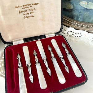 May include: A set of six antique mother-of-pearl handled knives in a black case with a red velvet interior. The knives have silver-toned metal accents and are arranged neatly. The case is labeled "Made in Sheffield, England expressly for Spencer Orgell, Beverly Hills, California."