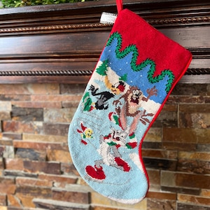 May include: A Christmas stocking with a red cuff and trim, featuring a cartoon scene with Bugs Bunny and other Looney Tunes characters. The stocking is hanging on a dark wooden mantelpiece, with a green wreath above.