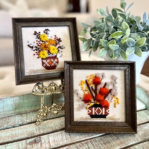 May include: Two framed cross-stitch embroidery pieces with fall-themed floral arrangements in brown pots. The pieces are displayed on a gold metal easel on a light blue wooden surface.