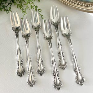May include: A set of seven antique silver forks with ornate floral and scroll detailing. The forks are arranged on a white surface, with a painting in the background. The forks are approximately 7 inches long.