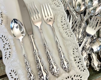 May include: A collection of ornate silverware, including spoons, knives, and forks, arranged on a white, decorative serving tray. The silverware has detailed, curved handles and a shiny, reflective surface. Additional silverware is visible in the background.