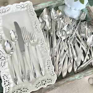 May include: A white tray with a decorative edge holds a set of silverware, including a knife, fork, and spoon. A wooden tray filled with a large collection of silver spoons and forks sits next to the white tray.