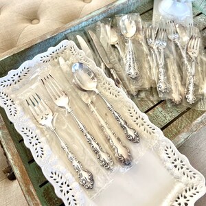 May include: A white ceramic tray with a lace pattern design, holding a set of silver flatware, including forks, spoons, and knives. The flatware is wrapped in clear plastic and arranged neatly on the tray.