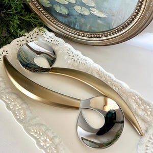 May include: Two serving spoons with a gold-colored handle and a silver-colored spoon bowl rest on a white, decorative tray. An oval painting in a silver frame is in the background. The painting depicts lily pads on a pond.