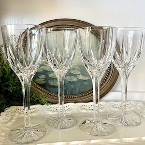 May include: Four clear crystal wine glasses with intricate cut glass designs. The glasses have long stems and circular bases. The background includes a framed painting and greenery, suggesting a vintage or elegant aesthetic.