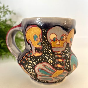 May include: A ceramic mug with a purple glaze and a black, white, yellow, and blue design featuring three skulls. The skulls have yellow faces with black eyes and black details. The mug has a handle.