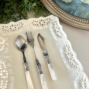 May include: A silver spoon, fork, and knife set with white handles. The utensils are arranged on a white, decorative plate. The handles have a decorative silver band. The set is likely for serving or eating.