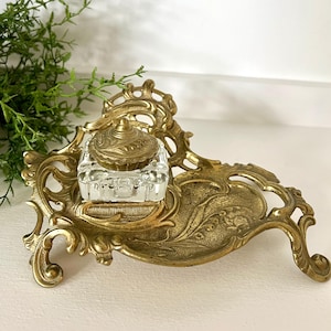 May include: An antique brass inkwell set with a clear glass ink pot and a decorative lid. The brass base features ornate scrollwork and a built-in pen rest. A vintage desk accessory.