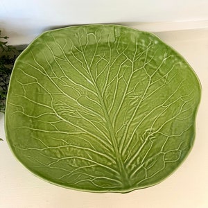 May include: A large, green ceramic serving dish shaped like a cabbage leaf. The dish has a detailed, textured design that resembles the veins of a cabbage leaf.