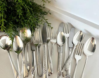 May include: An assortment of vintage silverware, including spoons, forks, and knives, arranged on a white surface. The silverware has ornate floral and scroll designs. Green foliage is in the background.