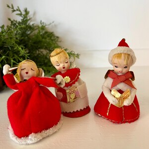 May include: Three vintage Christmas angel ornaments with blonde hair, red dresses, and gold halos. The angels are holding books with the word "Noel" printed on them.