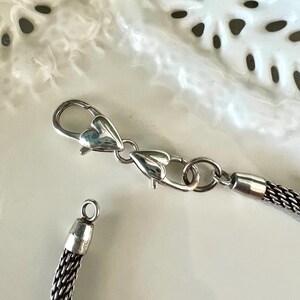 May include: Close-up of a silver necklace with a woven chain design. The necklace has a heart-shaped clasp and a small loop at the end. The chain is a dark silver colour, and the clasp is shiny.