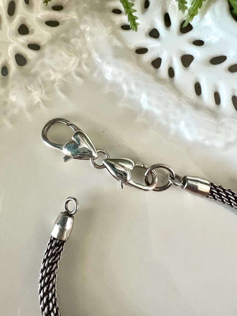 May include: Close-up of a silver necklace with a woven chain design. The necklace has a heart-shaped clasp and a small loop at the end. The chain is a dark silver colour, and the clasp is shiny.