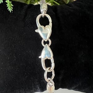 May include: A silver bracelet extender featuring heart-shaped clasps and decorative end pieces. The extender is designed to lengthen a bracelet or necklace. The clasps are heart-shaped, and the end pieces have a textured design.