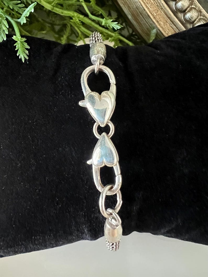 May include: A silver bracelet extender featuring heart-shaped clasps and decorative end pieces. The extender is designed to lengthen a bracelet or necklace. The clasps are heart-shaped, and the end pieces have a textured design.