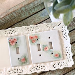 May include: Two white rectangular light switch plates featuring hand-painted pink rose clusters with green foliage. One plate is a single toggle switch cover, and the other is a double toggle switch cover, both displayed on a white ceramic tray.