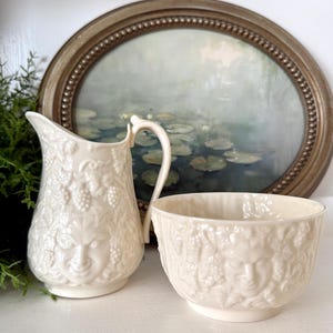 May include: Cream-colored ceramic pitcher and bowl set. The pitcher has a curved handle and is decorated with a face and grape vine design. The bowl has a similar design. An oval framed painting is in the background.