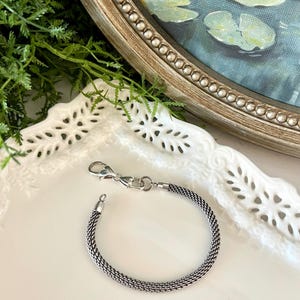 May include: A silver bracelet with a woven chain pattern and a lobster clasp. The bracelet is presented on a white, decorative plate. A painting of water lilies in a pond is visible in the background.