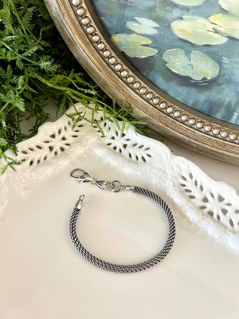 May include: A silver bracelet with a woven chain pattern and a lobster clasp. The bracelet is presented on a white, decorative plate. A painting of water lilies in a pond is visible in the background.