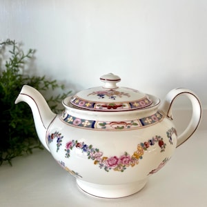 May include: White porcelain teapot with a floral design. The teapot features a lid and handle with a decorative floral garland around the body. The floral pattern includes pink, yellow, and blue flowers. The rim and handle are trimmed with a brown accent.