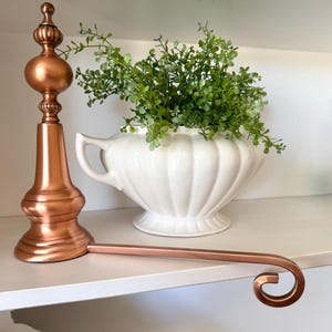 May include: A copper-colored metal stocking holder featuring a tall, decorative finial post with a rounded base and a long, horizontal arm ending in a curved hook. Behind the holder sits a white, fluted ceramic bowl filled with green foliage.