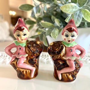 May include: A pair of vintage ceramic salt and pepper shakers. Each shaker is a whimsical elf figurine with a pink outfit, green collar, and a pointed red hat. The elves are perched on a brown tree stump base.