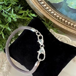 May include: A silver mesh bracelet with heart-shaped clasps is shown on a black velvet surface. The bracelet's woven design is visible, and the clasp is open. The background includes greenery and a framed painting.