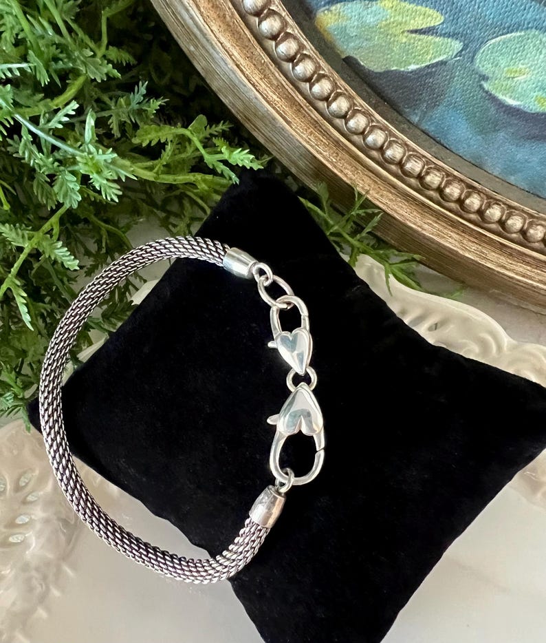 May include: A silver mesh bracelet with heart-shaped clasps is shown on a black velvet surface. The bracelet's woven design is visible, and the clasp is open. The background includes greenery and a framed painting.