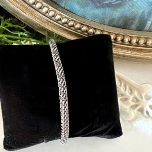 May include: A silver bracelet with a woven chain design is displayed on a black velvet cushion. The bracelet's texture and shine are highlighted against a backdrop of a framed painting and decorative elements.
