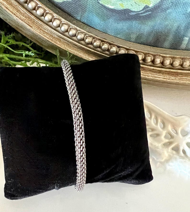 May include: A silver bracelet with a woven chain design is displayed on a black velvet cushion. The bracelet's texture and shine are highlighted against a backdrop of a framed painting and decorative elements.