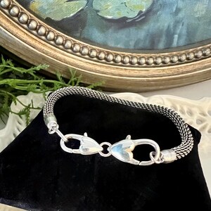 May include: A silver mesh bracelet with heart-shaped clasps. The bracelet is displayed on a black velvet surface, with a painting and ornate gold frame in the background. The bracelet's clasp is a unique design.