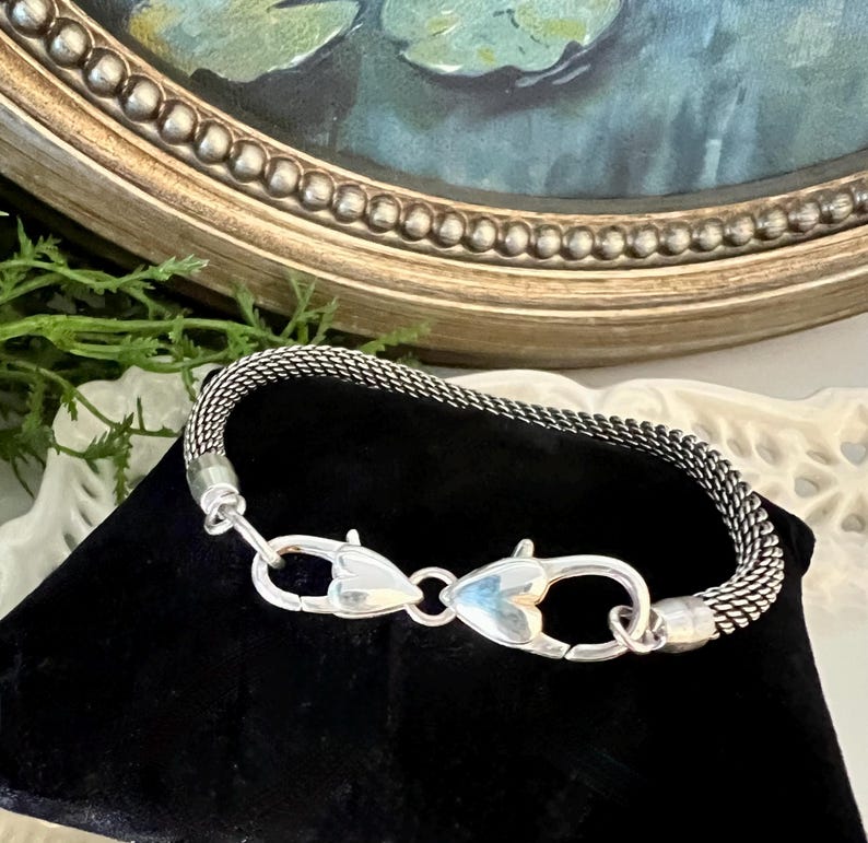 May include: A silver mesh bracelet with heart-shaped clasps. The bracelet is displayed on a black velvet surface, with a painting and ornate gold frame in the background. The bracelet's clasp is a unique design.
