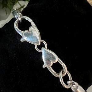 May include: A silver bracelet with heart-shaped clasps. The bracelet is made of polished metal and features two heart-shaped clasps that connect the chain. The bracelet is displayed on a black velvet surface.