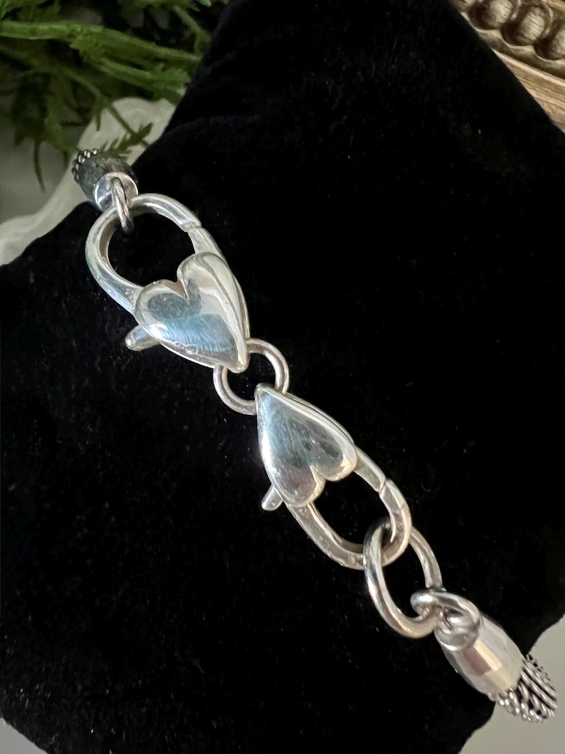 May include: A silver bracelet with heart-shaped clasps. The bracelet is made of polished metal and features two heart-shaped clasps that connect the chain. The bracelet is displayed on a black velvet surface.
