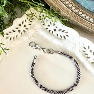 May include: A silver bracelet with a woven chain pattern. It has a lobster clasp fastening and a small loop at the other end. The bracelet is presented on a white, decorative plate.