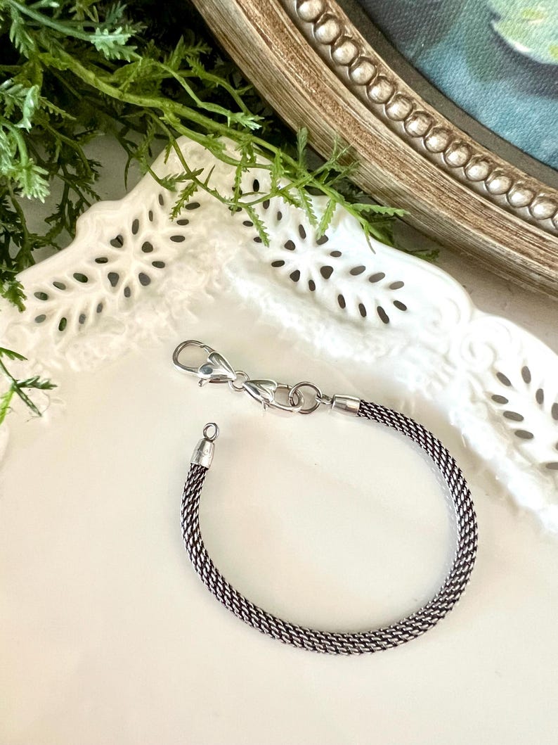 May include: A silver bracelet with a woven chain pattern. It has a lobster clasp fastening and a small loop at the other end. The bracelet is presented on a white, decorative plate.